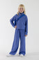 A full front view of a young girl with blonde hair smiling and standing hand-on-hip, wearing a bluish-purple ribbed quarter-zip pullover, matching wide-leg pants, and white sneakers, styled with a matching Perla Blue belt bag slung across her chest against a plain light background.