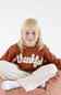 A young blonde girl sitting cross-legged on a dusty rose beanbag chair, wearing a rust-brown sweatshirt with the word 'thankful' in white script across the chest, paired with cream-colored sweatpants.