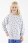 A young blonde girl smiling while wearing a light gray long-sleeve sweatshirt with a black polka dot pattern, paired with blue pants, against a plain white background.