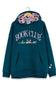 A dark teal blue hoodie with a pink polka-dot lined hood, featuring 'BOOK CLUB Since 1883' printed in pink and white on the front. The hoodie is displayed on a wooden hanger against a plain white background.