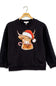 A black crewneck sweatshirt hanging on a wooden hanger, featuring a graphic of a Highland cow wearing a red Santa hat and holding a candy cane.