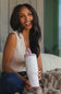 A woman smiling while holding a tall insulated water bottle with a pink lid and a white body covered in small pink floral speckles, seated indoors on a couch with a blanket.