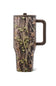 A travel tumbler with a woodland camouflage pattern featuring brown branches, green leaves, and beige bark, with a dark brown handle and lid, shown on a white background.