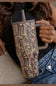A close-up of a person holding a tall handled tumbler with a realistic woodland camouflage pattern. The tumbler has a gray handle and lid, and the person is wearing a black top and blue denim jeans.
