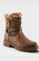 A brown leather ankle boot with a faux-fur collar, a decorative strap with studs and a buckle around the ankle, and a low block heel, shown on a light gray background.
