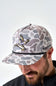 A man wearing a camouflage baseball cap with a flying duck graphic and a black rope detail under the brim. The cap features a light gray and brown camo pattern, and the brand name 'BURLEBO' is visible on the side.