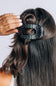 A close-up of a woman's hair styled with a half-up bun, wearing a black hair claw clip with white polka dots. A hand with white manicured nails is adjusting the clip.