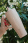 A hand with red nail polish holds a tall, tapered travel tumbler with a handle and straw. The tumbler has a light pink and white vertical stripe pattern and features the brand name 'BRuMATE' printed near the bottom.