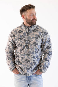 A man wearing a light gray marled quarter-zip knit sweater and dark jeans, standing against a plain white studio background.