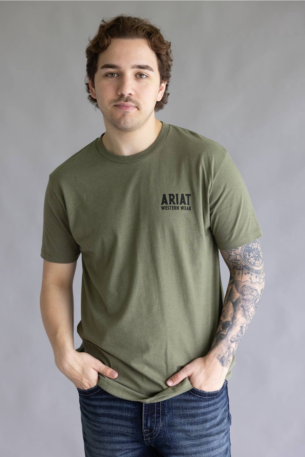 A man wearing an olive green crewneck t-shirt with 'ARIAT WESTERN WEAR' printed in black on the left chest, paired with blue jeans, standing against a neutral gray background.