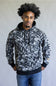 A man wearing a black and white abstract patterned hoodie with black drawstrings, black ribbed cuffs and hem, paired with blue jeans, standing against a plain light gray background.