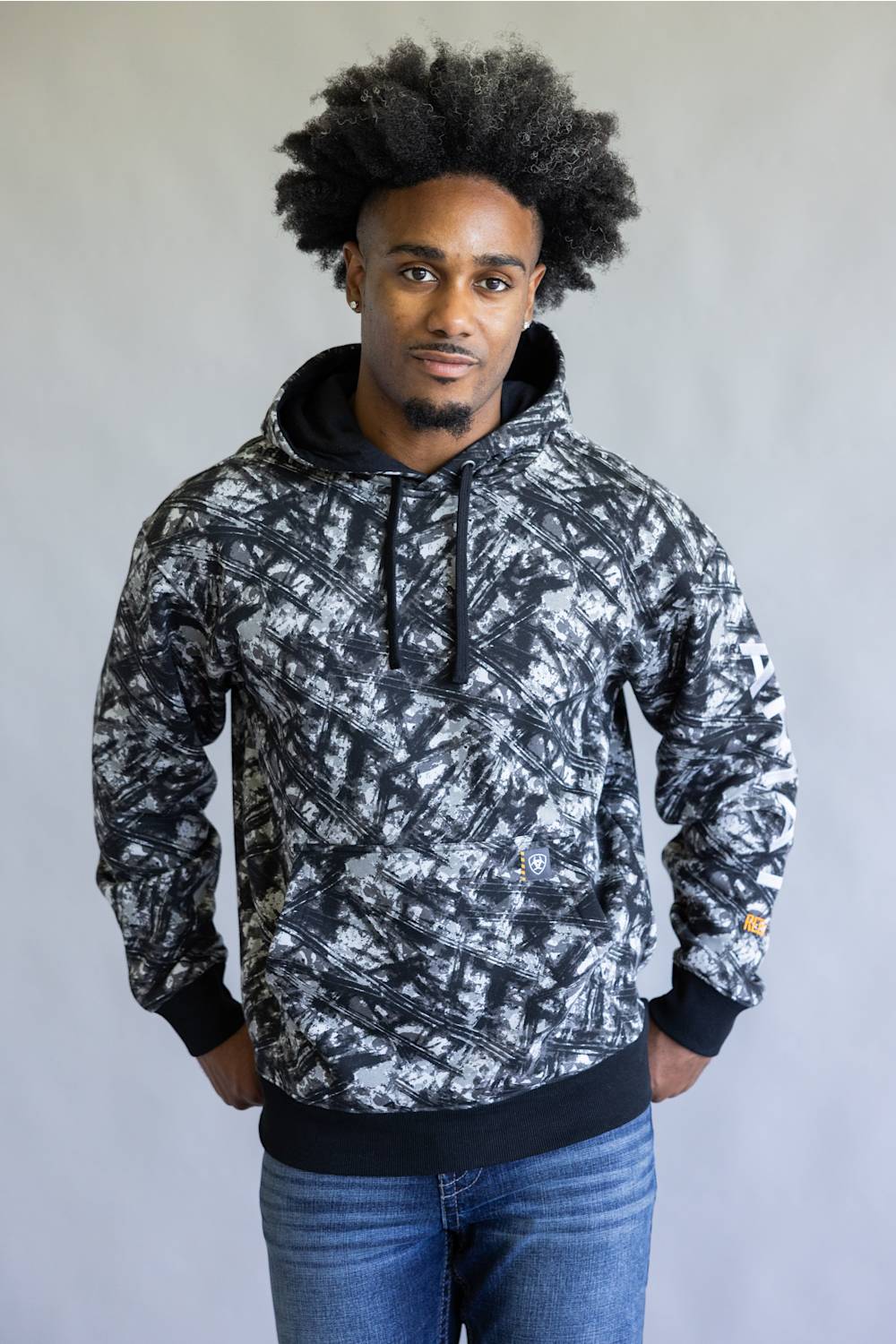 A man wearing a black and white abstract patterned hoodie with black drawstrings, black ribbed cuffs and hem, paired with blue jeans, standing against a plain light gray background.
