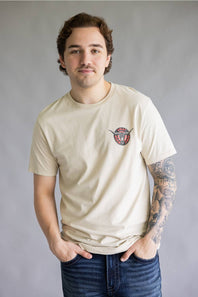 A man wearing a beige crewneck t-shirt with a small circular graphic on the left chest, paired with dark blue jeans, standing against a neutral gray background.