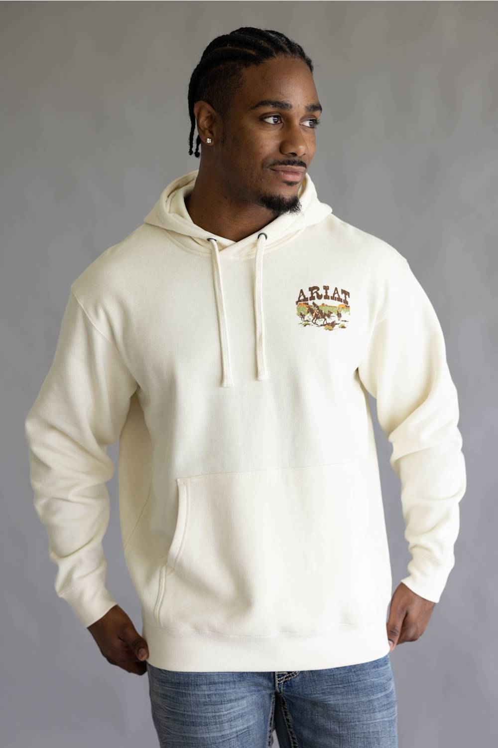 A man wearing a cream-colored hoodie with a drawstring hood and a kangaroo pocket, featuring a small graphic on the left chest that reads 'ARIAN' with a horse and rider illustration. He is also wearing blue jeans, standing against a neutral gray studio background.