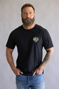 Front view of a bearded man with tattooed arms posing in a black short-sleeve t-shirt featuring a small white Ariat logo on the left chest, paired with blue jeans.