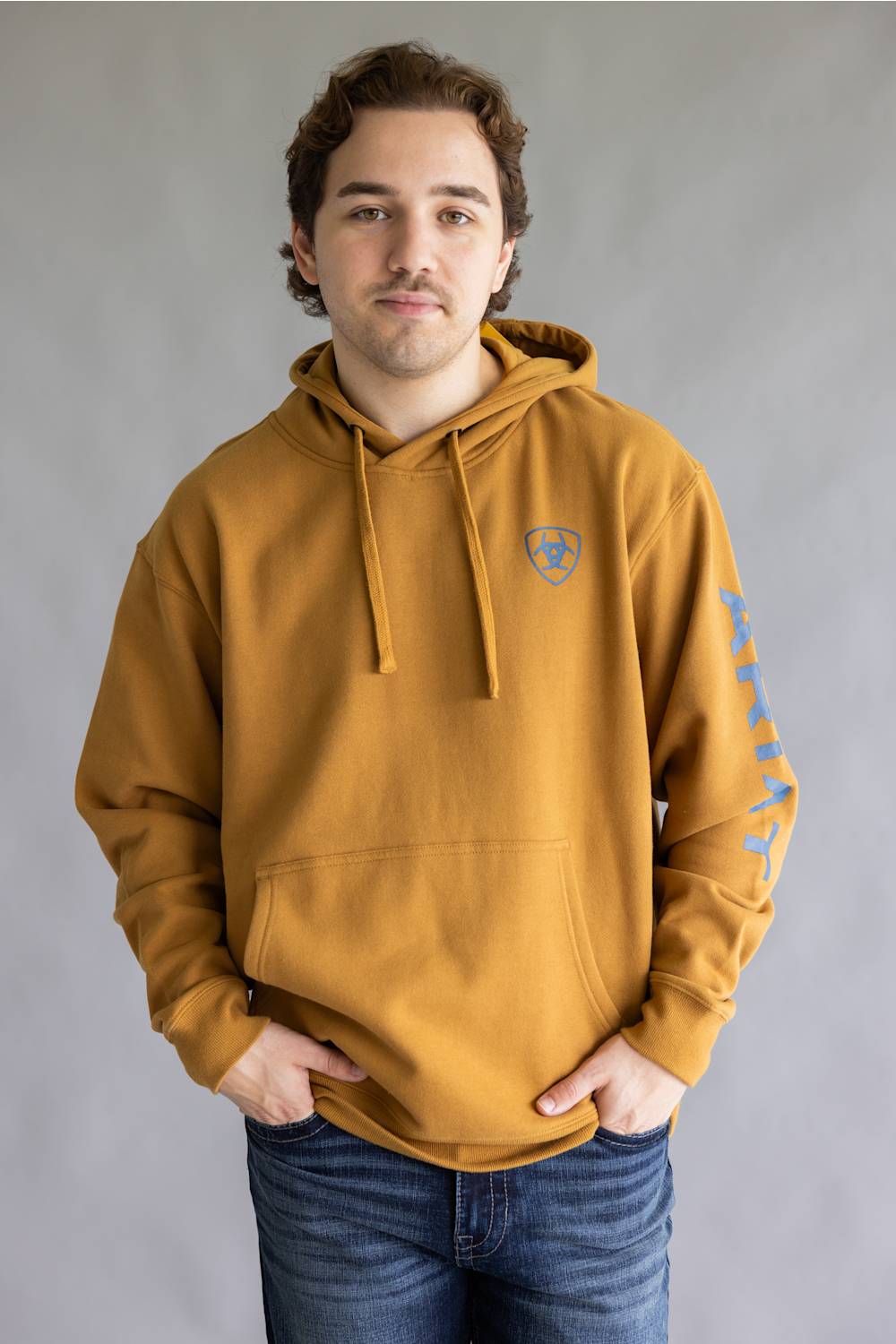 A man wearing a mustard yellow hoodie with a blue shield logo on the left chest and blue graphics on the right sleeve, paired with blue jeans, standing against a neutral gray background.