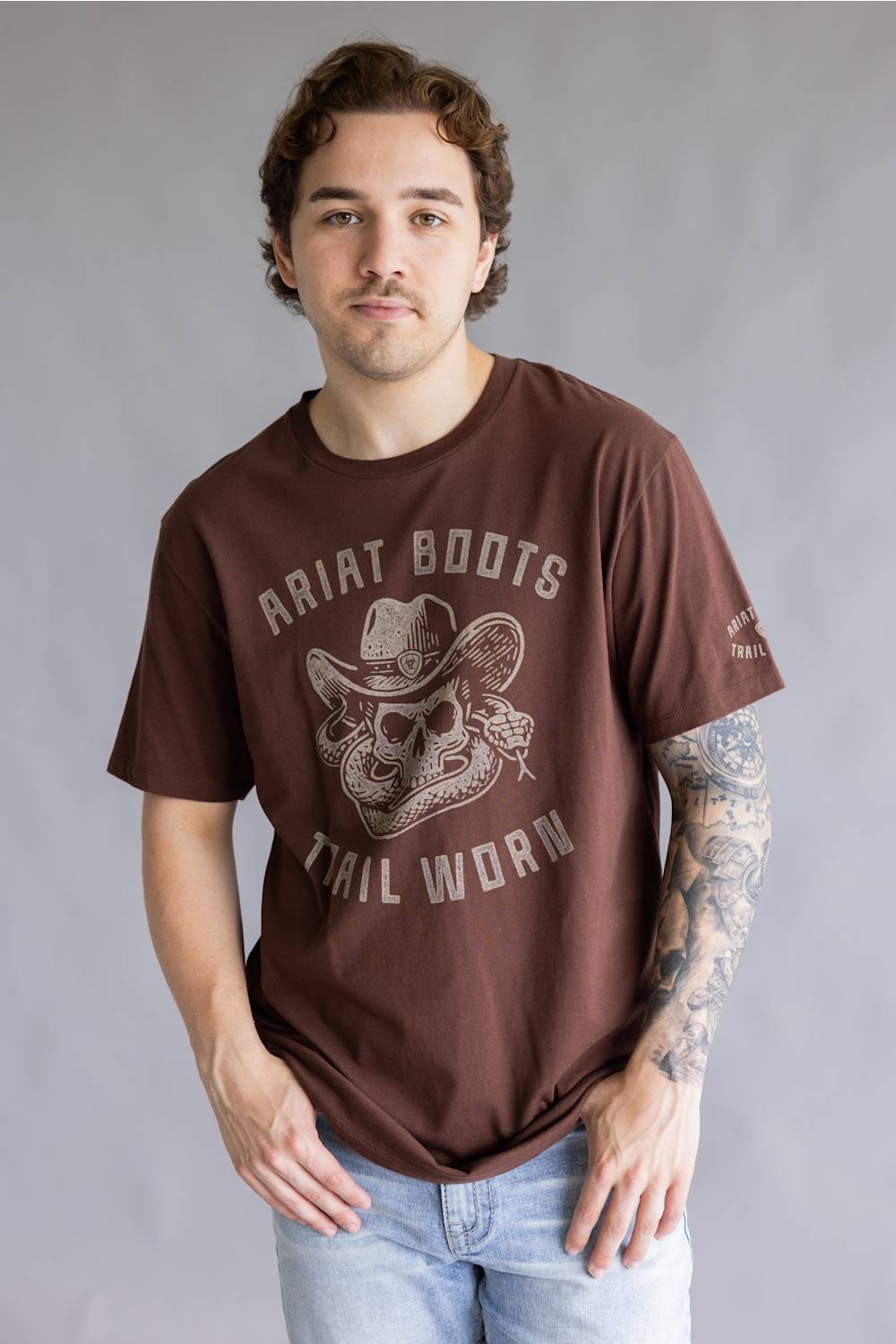 A man wearing a brown graphic t-shirt with 'Ariat Boots Tail Worn' printed in distressed white lettering and a cowboy skull design, paired with light blue jeans against a gray studio background.