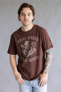 A man wearing a brown graphic t-shirt with 'Ariat Boots Tail Worn' printed in distressed white lettering and a cowboy skull design, paired with light blue jeans against a gray studio background.
