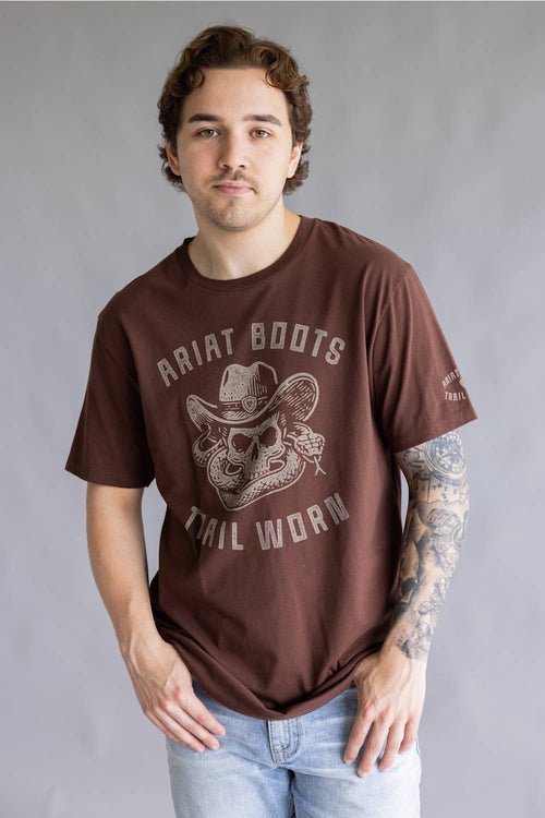 A man wearing a brown graphic t-shirt with 'Ariat Boots Tail Worn' printed in distressed white lettering and a cowboy skull design, paired with light blue jeans against a gray studio background.