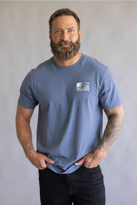 Front view of a bearded man with hands in his pockets wearing a slate blue short-sleeve t-shirt.