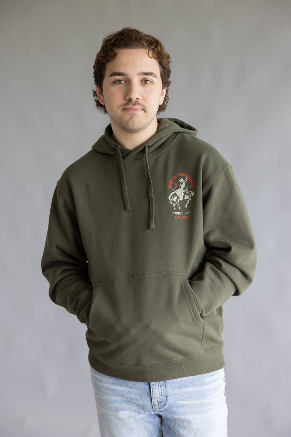A man wearing an olive green pullover hoodie with a graphic print on the left chest, paired with light blue jeans, standing against a neutral gray studio background.