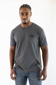 A man wearing a dark gray crewneck t-shirt with a small black chest logo that reads 'Ariat', paired with blue jeans, standing against a plain white background.