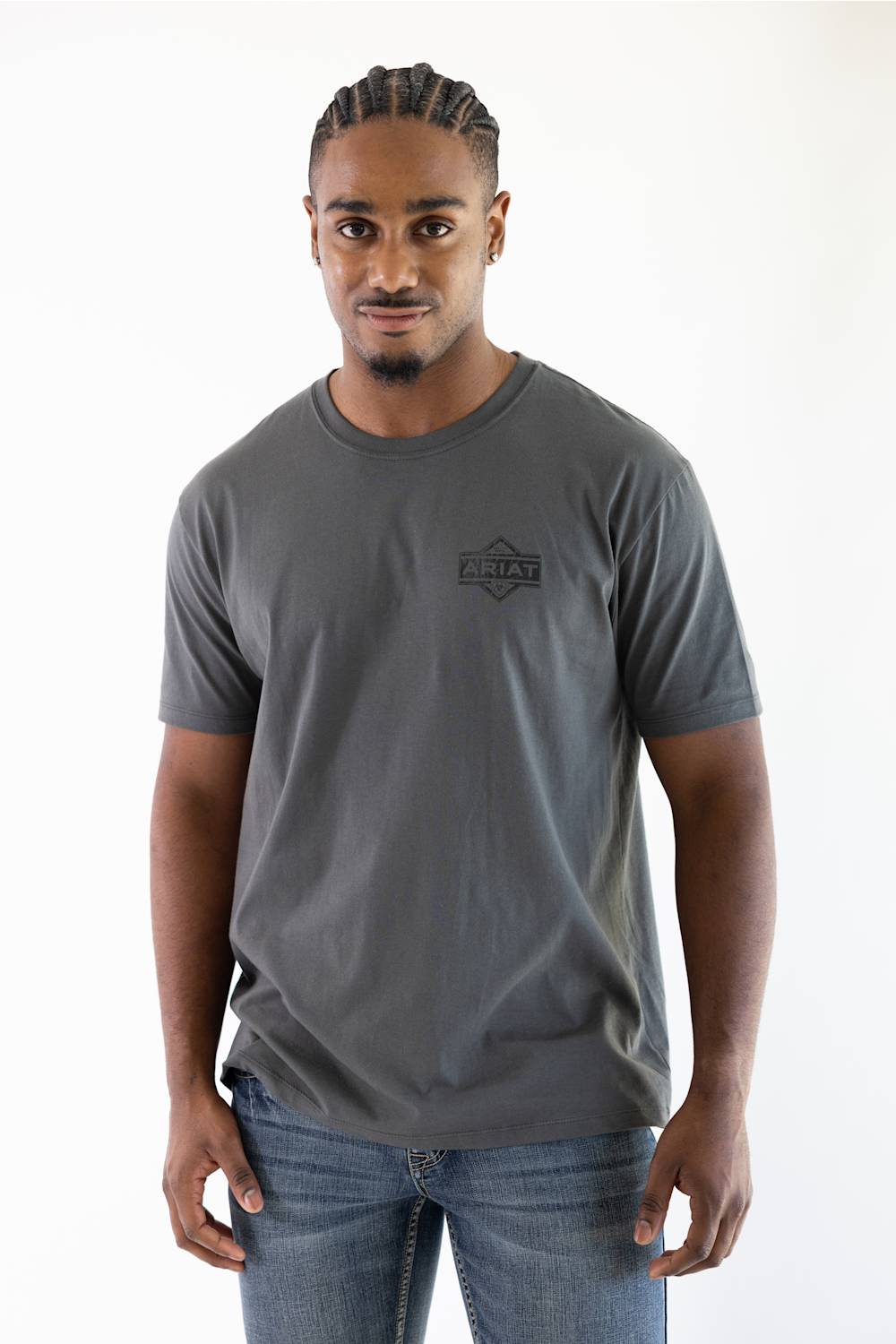 A man wearing a dark gray crewneck t-shirt with a small black chest logo that reads 'Ariat', paired with blue jeans, standing against a plain white background.