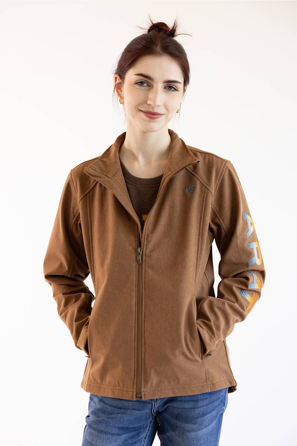 A woman wearing a brown zip-up jacket with a stand collar, side pockets, and a multi colored logo graphic print on the left sleeve, paired with a dark brown t-shirt and blue jeans, against a plain white background.