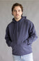 A man wearing a dark slate blue hoodie with a tonal embroidered logo on the left chest, paired with light blue jeans, standing against a neutral gray studio background.