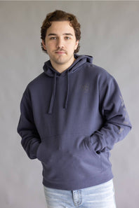 A man wearing a dark slate blue hoodie with a tonal embroidered logo on the left chest, paired with light blue jeans, standing against a neutral gray studio background.