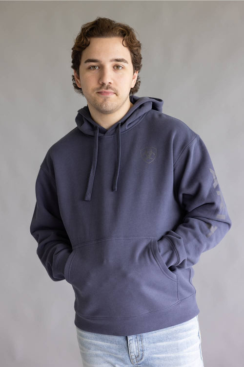 A man wearing a dark slate blue hoodie with a tonal embroidered logo on the left chest, paired with light blue jeans, standing against a neutral gray studio background.