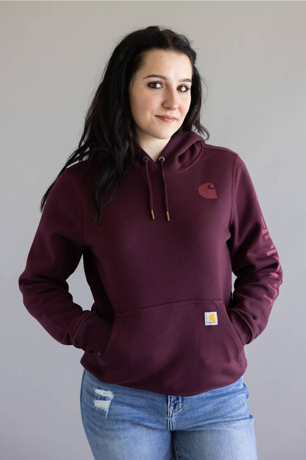 A woman wearing a maroon Carhartt hoodie with a small embroidered Carhartt logo on the left chest and a signature yellow square patch on the front pocket, paired with blue jeans against a plain gray background.