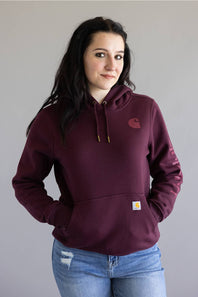 A woman wearing a maroon Carhartt hoodie with a small embroidered Carhartt logo on the left chest and a signature yellow square patch on the front pocket, paired with blue jeans against a plain gray background.