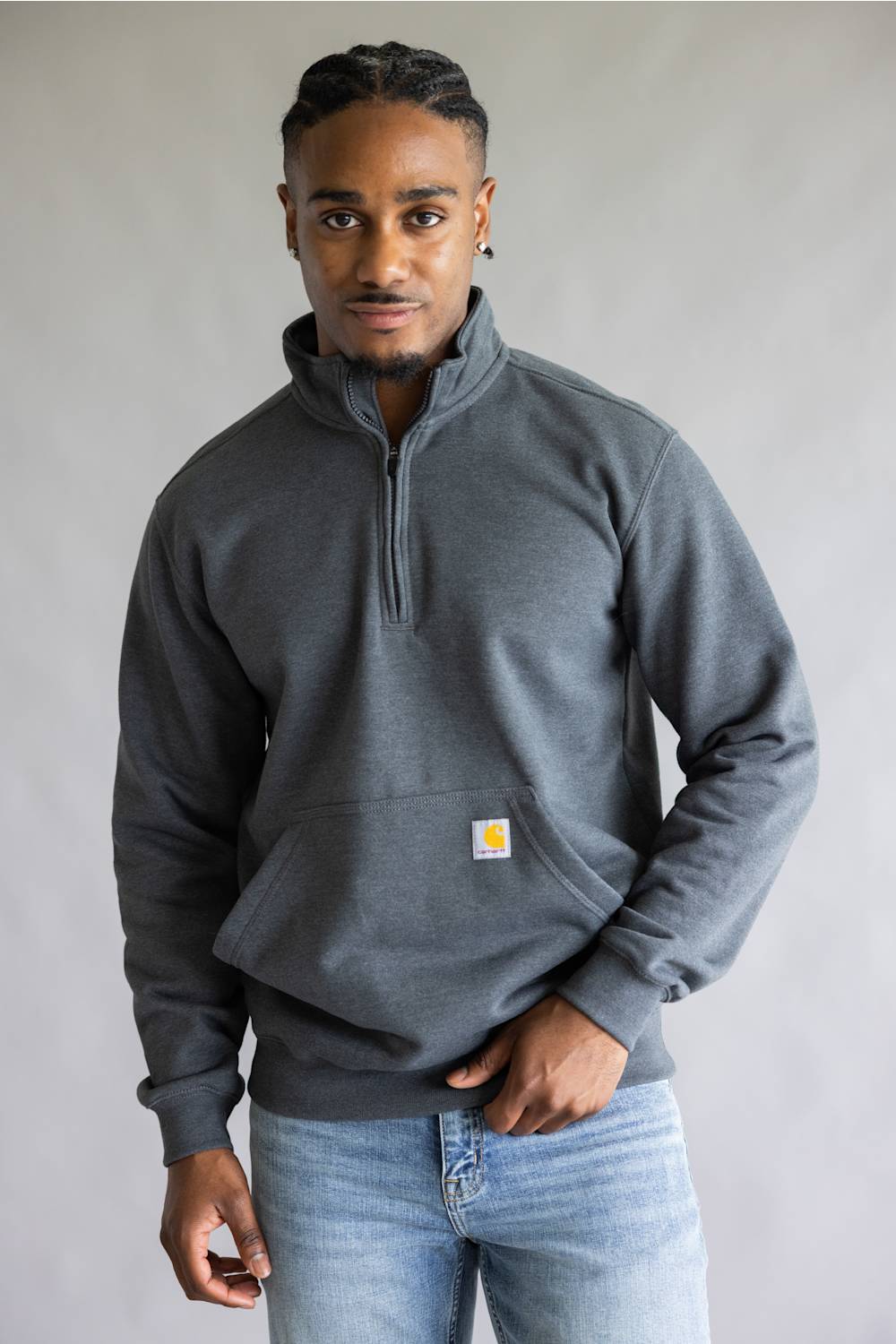 A man wearing a dark gray quarter-zip sweatshirt with a small Carhartt logo patch on the chest, paired with light blue jeans, standing against a neutral gray background.