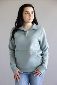 A woman wearing a light blue-gray quarter-zip sweatshirt with a small Carhartt logo patch on the front pocket, paired with blue jeans, standing against a plain gray background.
