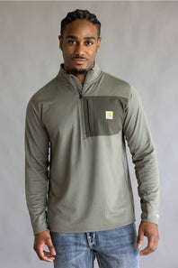 A man wearing a sage green ribbed quarter-zip pullover with a chest pocket and a small Carhartt logo patch, paired with blue jeans, against a neutral gray background.