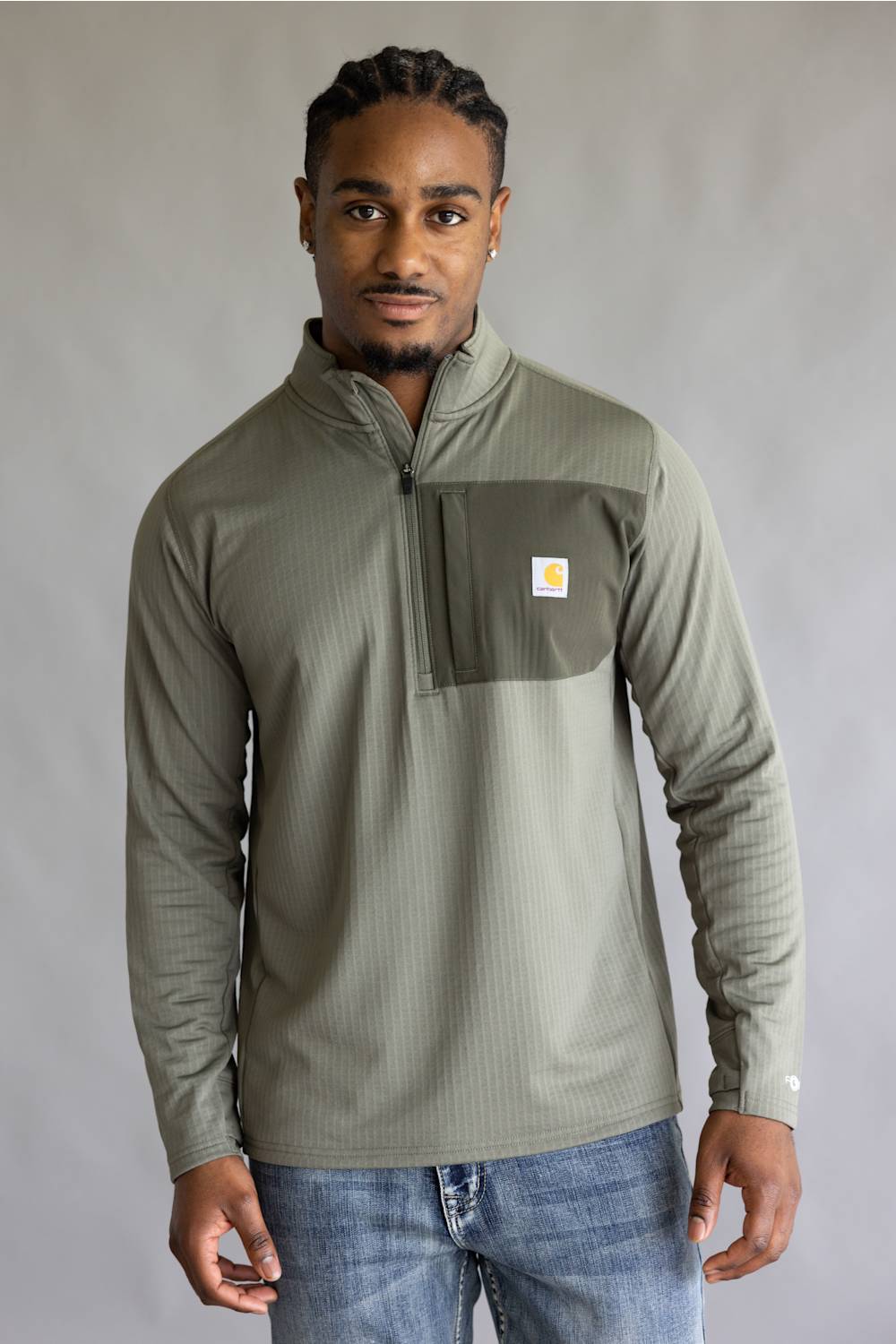 A man wearing a sage green ribbed quarter-zip pullover with a chest pocket and a small Carhartt logo patch, paired with blue jeans, against a neutral gray background.