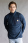 A man wearing a navy blue quarter-zip pullover sweatshirt with a small Carhartt logo patch on the left chest, paired with light blue jeans, standing against a neutral gray studio background.
