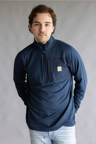 A man wearing a navy blue quarter-zip pullover sweatshirt with a small Carhartt logo patch on the left chest, paired with light blue jeans, standing against a neutral gray studio background.