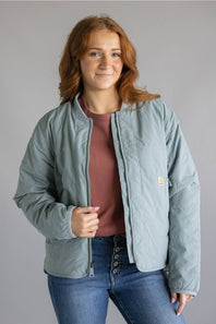 A woman wearing a light sage green quilted jacket by Carhartt, layered over a dusty rose top and blue jeans, standing against a solid gray studio background.