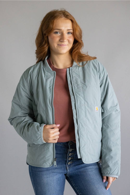 A woman wearing a light sage green quilted jacket by Carhartt, layered over a dusty rose top and blue jeans, standing against a solid gray studio background.