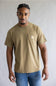 A man wearing a tan Carhartt short-sleeve crewneck t-shirt with a chest pocket and blue jeans, standing against a neutral gray background.