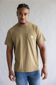 A man wearing a tan Carhartt short-sleeve crewneck t-shirt with a chest pocket and blue jeans, standing against a neutral gray background.
