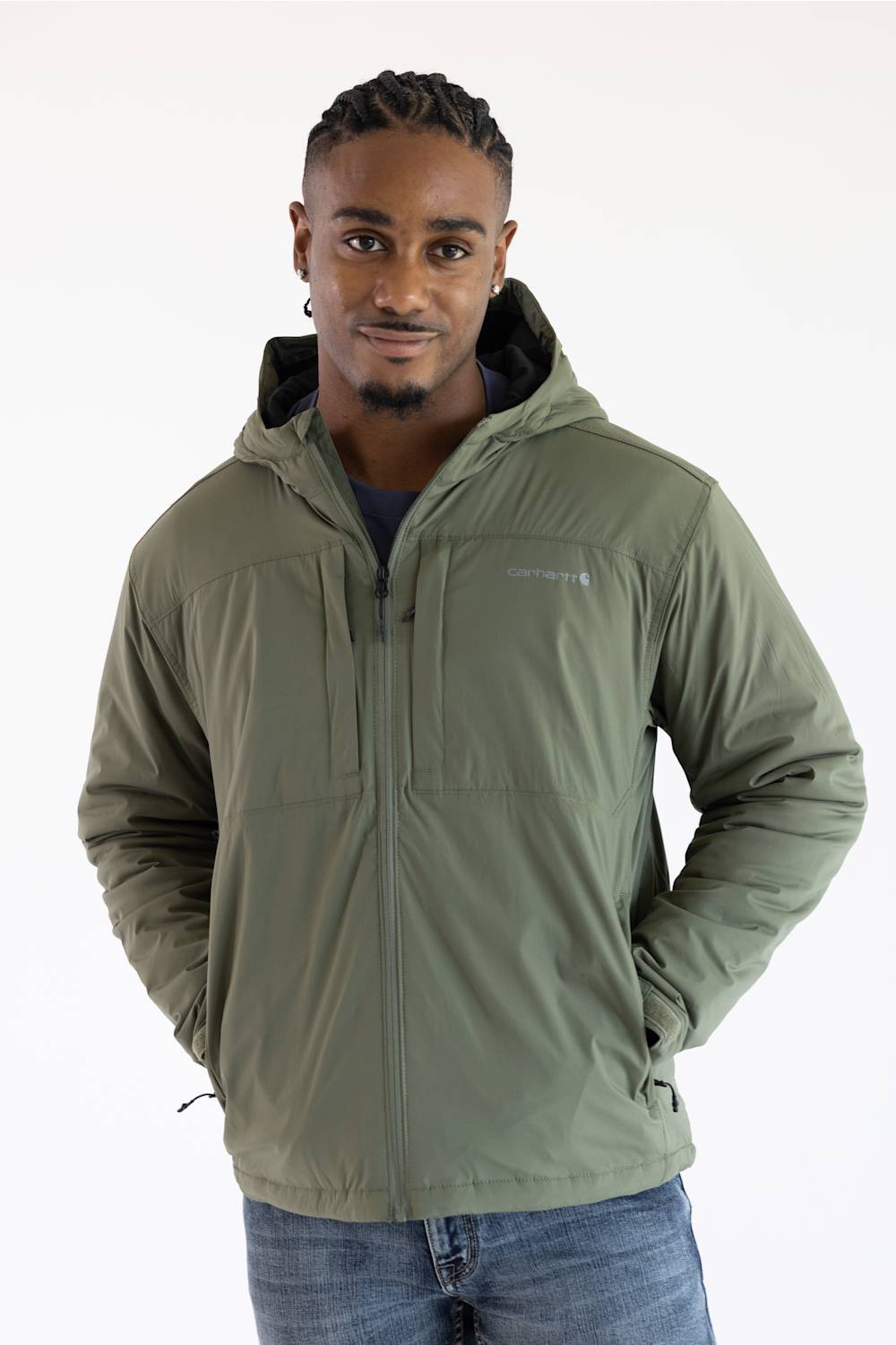 A man wearing a dusty olive green hooded Carhartt jacket paired with blue jeans, posed against a plain white background with hands in his pockets.