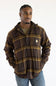 A man wearing a brown and gold plaid hooded button-up shirt jacket by Carhartt, paired with a green t-shirt and blue jeans, against a plain white background.