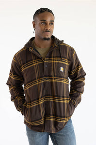 A man wearing a brown and gold plaid hooded button-up shirt jacket by Carhartt, paired with a green t-shirt and blue jeans, against a plain white background.