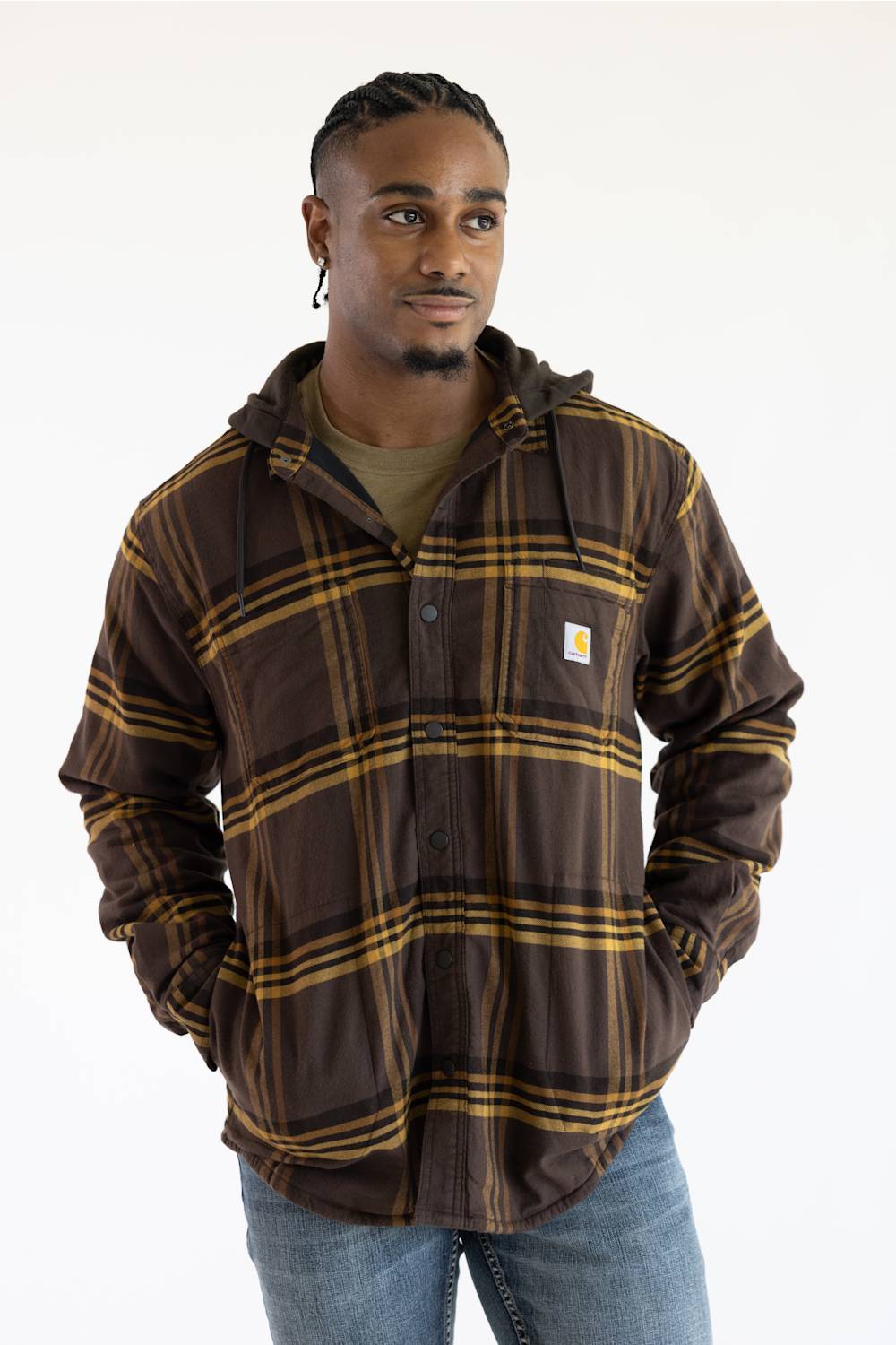 A man wearing a brown and gold plaid hooded button-up shirt jacket by Carhartt, paired with a green t-shirt and blue jeans, against a plain white background.