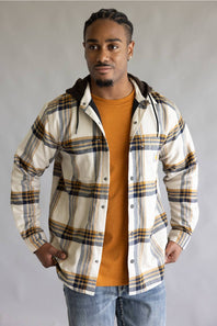 A man wearing a cream-colored hooded plaid overshirt with navy and mustard yellow stripes, layered over an orange t-shirt and blue jeans, standing against a neutral gray background.