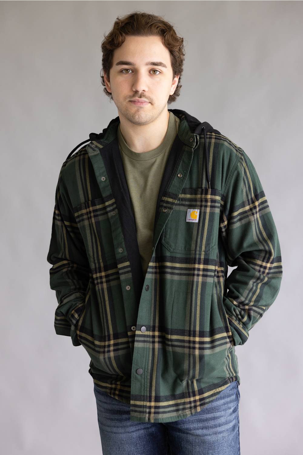 A man wearing a green Carhartt plaid hooded overshirt over an olive green t-shirt and blue jeans, standing against a neutral gray studio background.