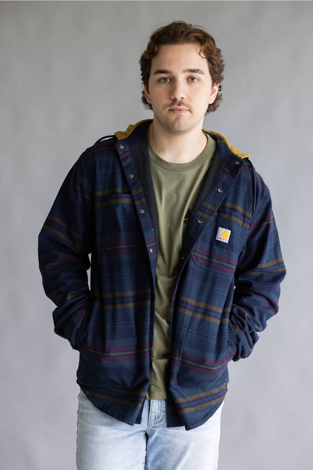 A man wearing a navy blue hooded plaid overshirt with yellow and red stripes, layered over an olive green t-shirt and light wash jeans, standing against a gray studio background.
